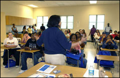 [Photo: View of a speaker addressing a large group of adults classroom with all desks occupied and eight people standing in the back]