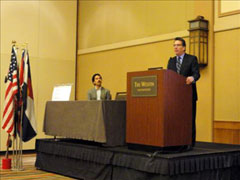[Photo 3: Rick Garcia speaking at podium with Randy Akers listening in background]